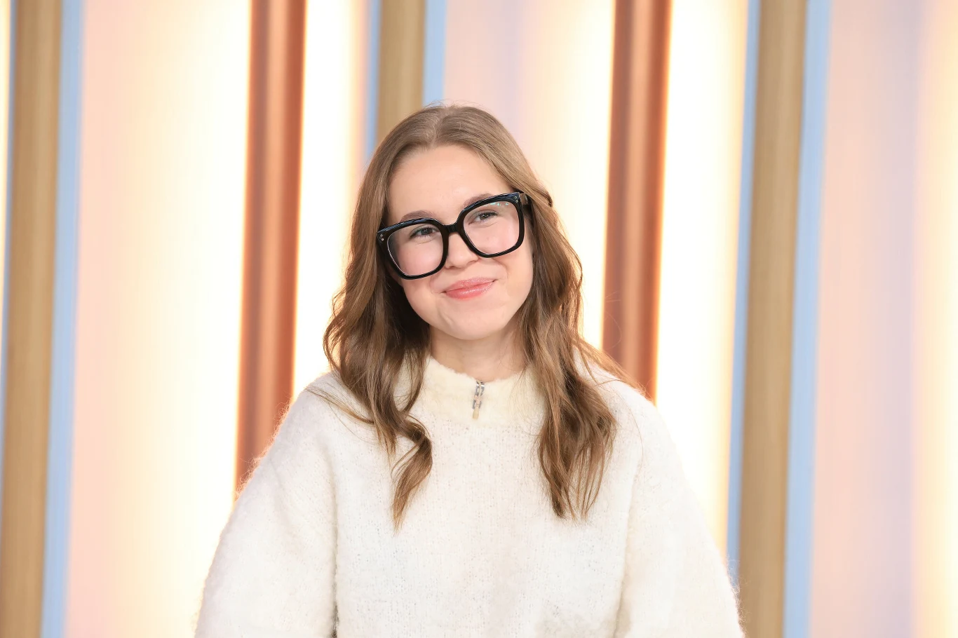 Zoja Skubis Młoda kobieta z długimi, jasnobrązowymi włosami i dużymi okularami w czarnej oprawie, uśmiechnięta, ubrana w jasny, miękki sweter, na tle kolorowych pionowych paneli.