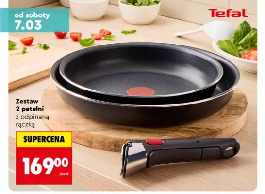 Dwie czarne patelnie firmy Tefal ustawione jedna w drugiej na drewnianej desce kuchennej, obok zdejmowana rączka, w tle widoczne świeże warzywa i zioła na kuchennym blacie.