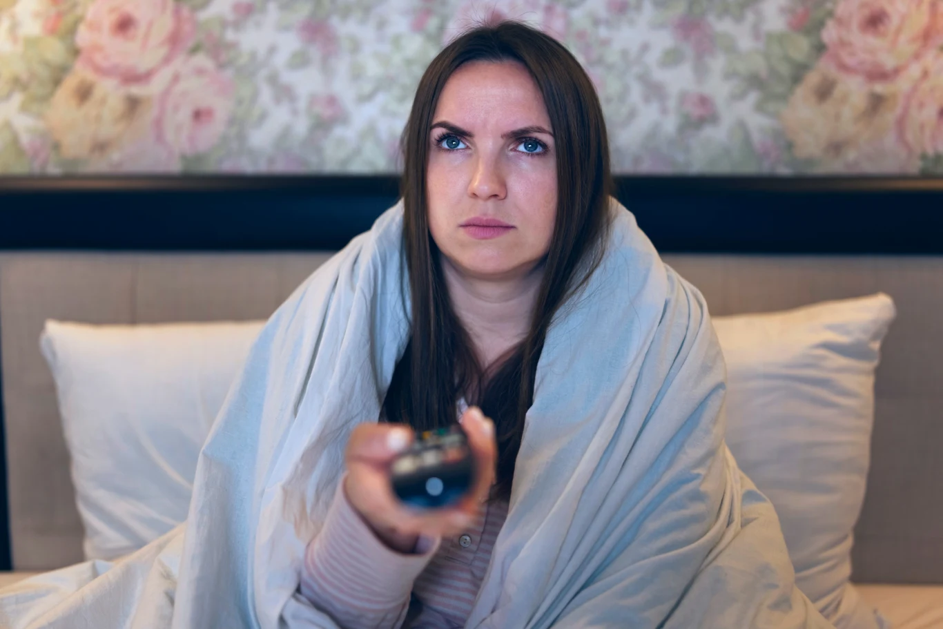 Kobieta siedzi na łóżku owinięta kocem, trzyma w ręce pilot do telewizora i patrzy przed siebie z poważnym wyrazem twarzy.