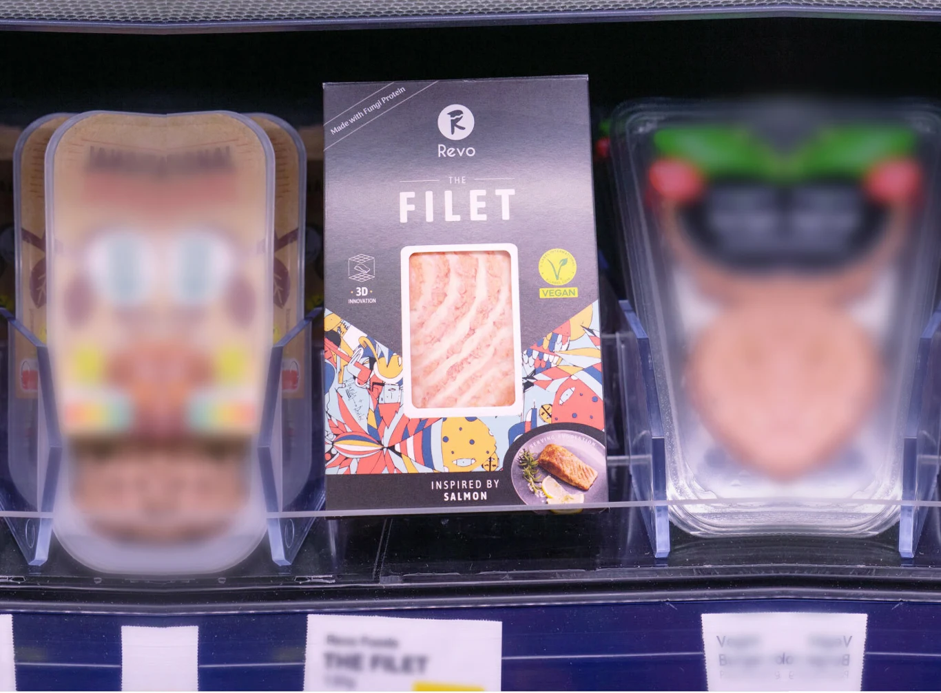 Roślinny filet inspirowany łososiem w ciemnym opakowaniu z kolorową grafiką, wyeksponowany na półce chłodniczej pomiędzy innymi produktami imitującymi mięso o rozmazanych etykietach.