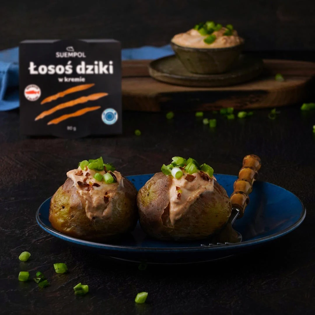 Dwa pieczone ziemniaki podane na niebieskim talerzu, nadziewane kremem z dzikiego łososia i posypane świeżą posiekaną dymką, z boku widoczny widelec, w tle opakowanie kremu z łososia oraz miseczka z dodatkiem kremu i dymką.