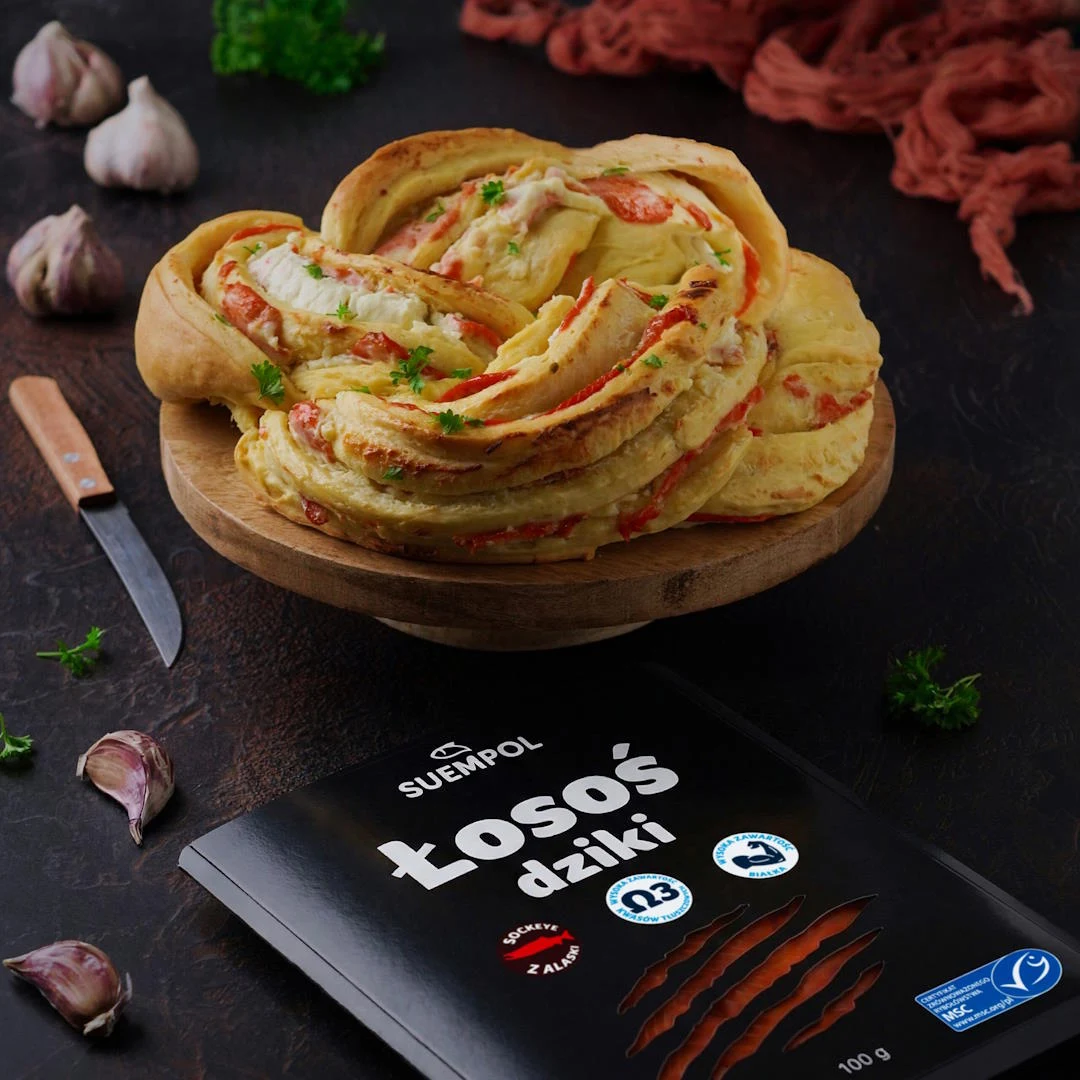 Na drewnianej misie znajduje się efektownie uformowana drożdżowa bułka z widocznymi kawałkami łososia i zieleniną, obok leży opakowanie dzikiego łososia Suempol, wokół znajdują się ząbki czosnku, nóż i świeże zioła.