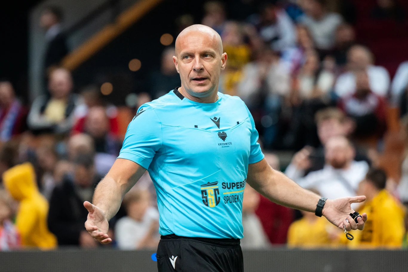 Łysy sędzia w niebieskim stroju sportowym stoi na boisku z rozłożonymi rękami, w tle widownia na trybunach.