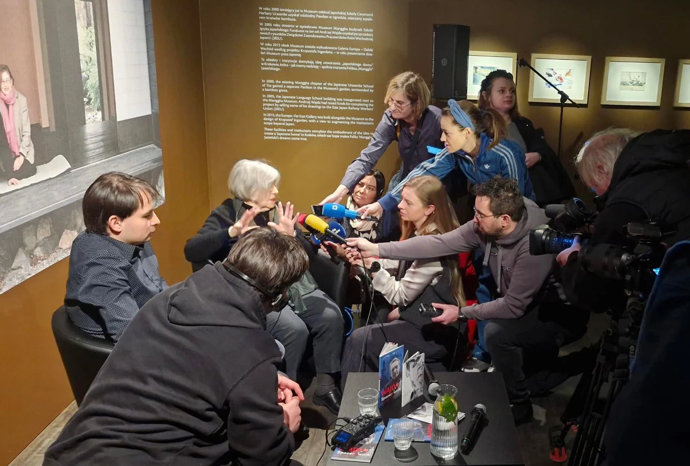 Starsza kobieta siedząca naprzeciwko grupy dziennikarzy przekazujących jej mikrofony, obok niej młodszy mężczyzna, w tle tablica informacyjna i zdjęcia na ścianie, atmosfera dynamiczna, skupienie na rozmowie i rejestrowaniu wypowiedzi.