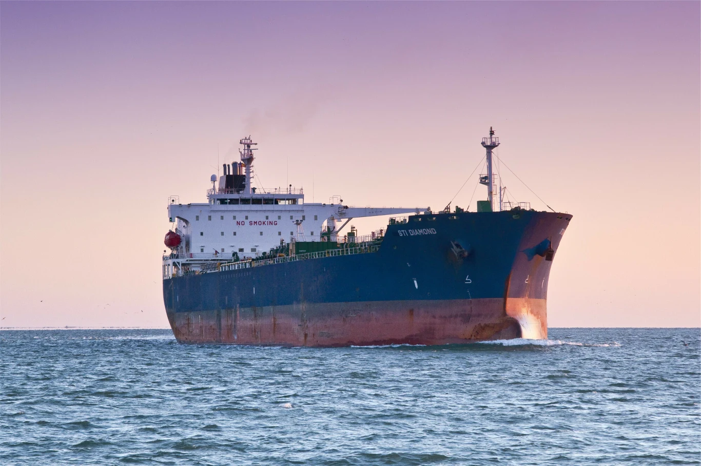 Duży tankowiec płynący po spokojnym morzu podczas zachodu słońca, z widoczną ciemnoniebieską burtą i jasno oznaczonym dziobem.