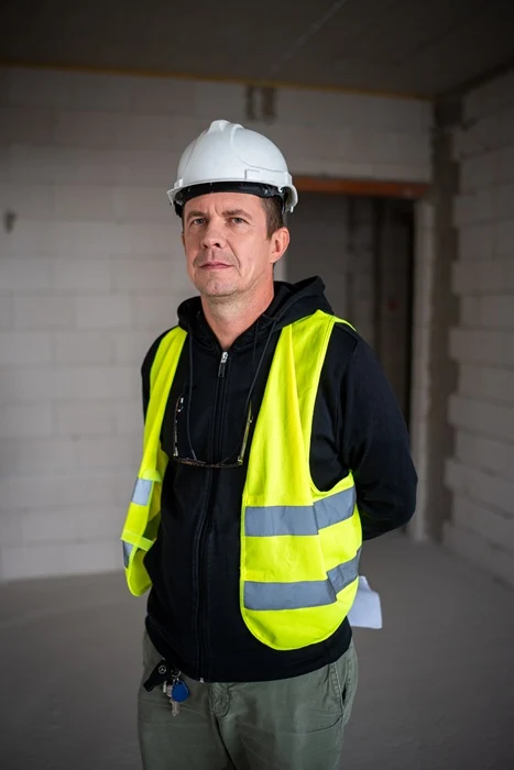 Mężczyzna w kamizelce odblaskowej i białym kasku ochronnym stoi wewnątrz budynku w trakcie budowy; ma założone okulary ochronne na szyi i ręce schowane za plecami.