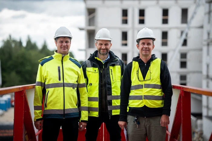 Trzech mężczyzn w kaskach ochronnych i odblaskowych kamizelkach stoi na placu budowy, w tle widoczny jest budynek w trakcie wznoszenia.