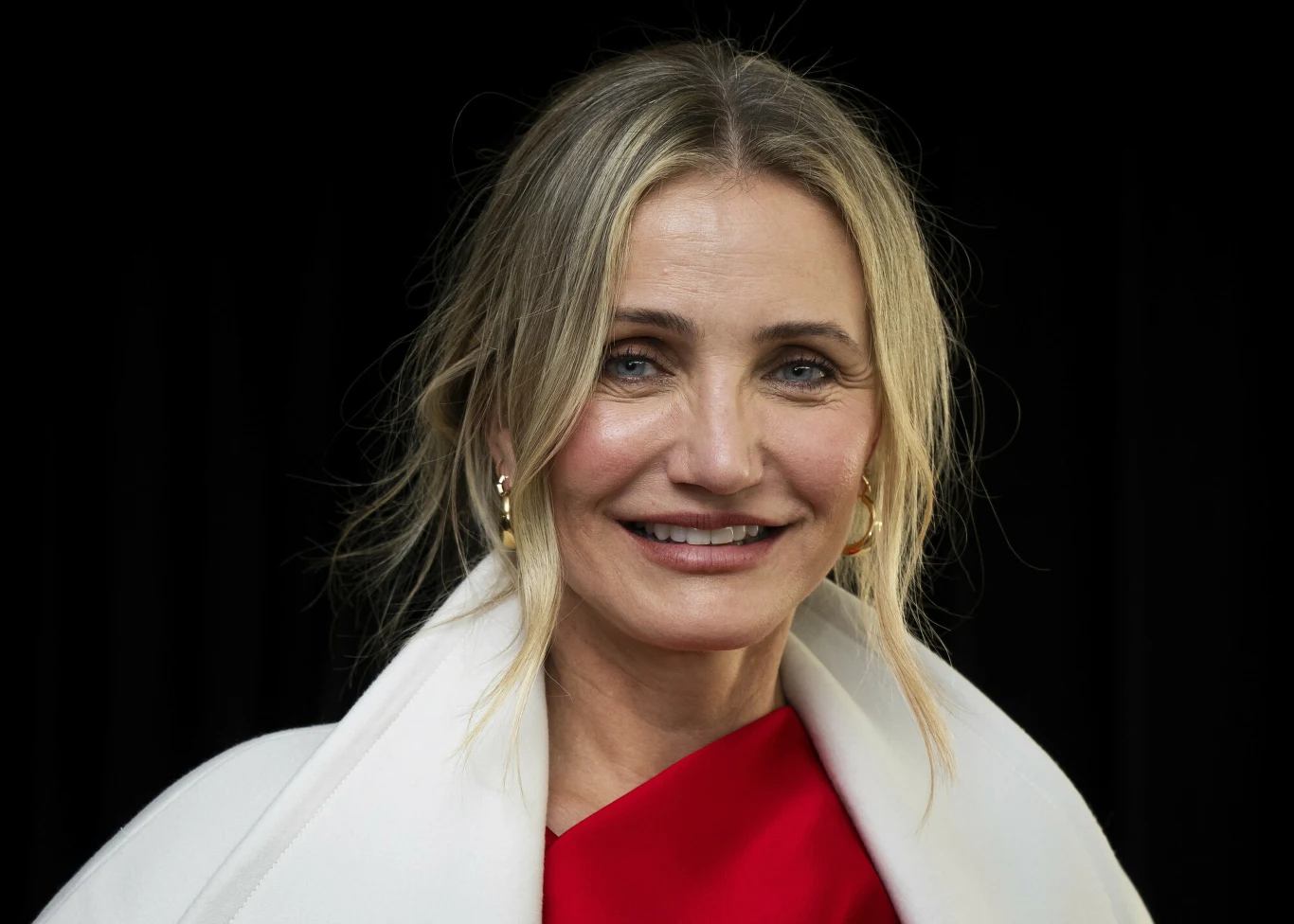 Kobieta z jasnymi włosami upiętymi w luźny kok, ubrana w elegancką białą pelerynę i czerwoną suknię, uśmiecha się patrząc prosto w obiektyw na tle czarnego tła.