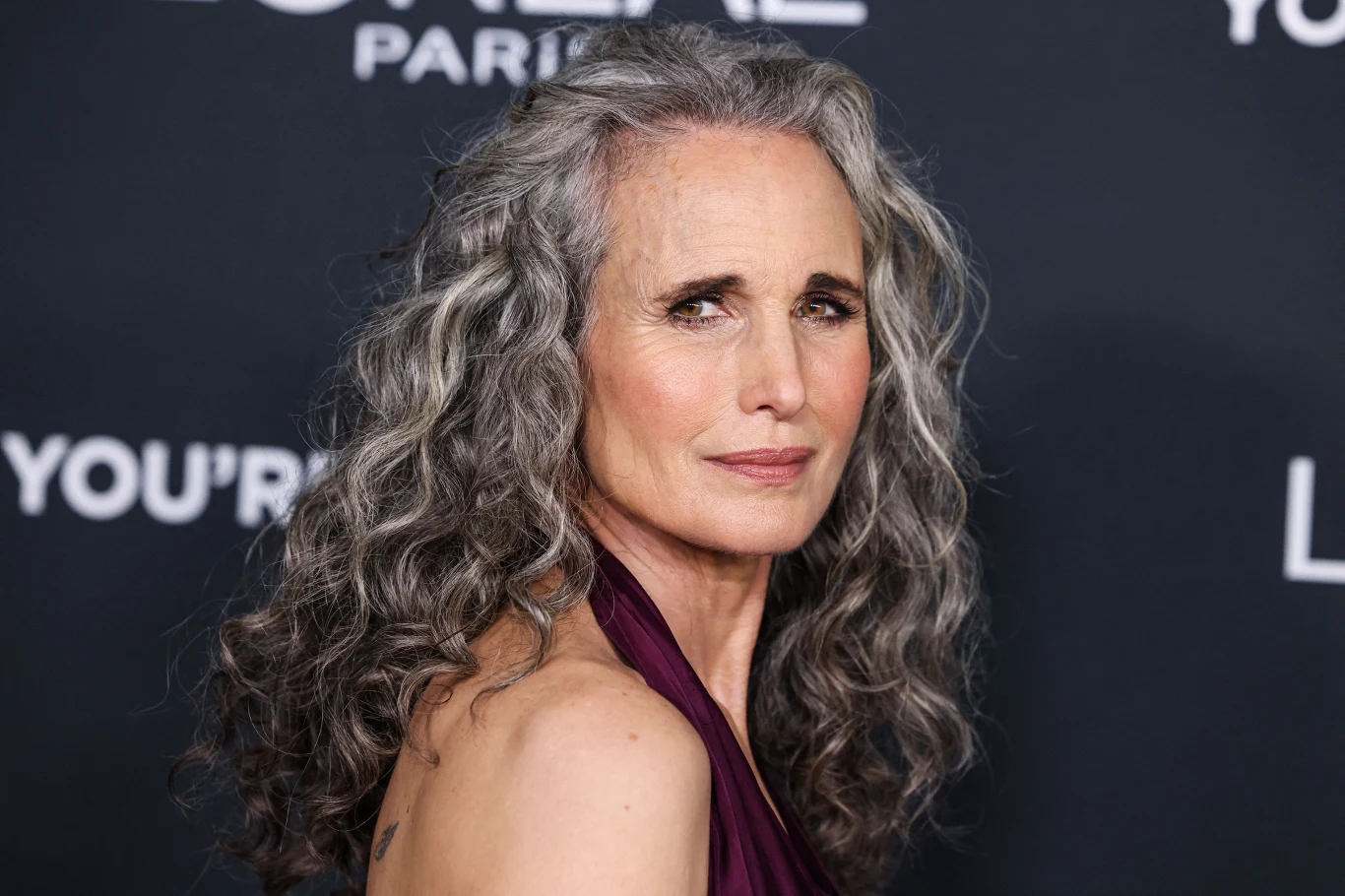 Kobieta o długich, siwych, falowanych włosach i naturalnym wyglądzie, ubrana w bordową suknię bez rękawów, patrząca poważnie w stronę kamery na tle ciemnej ścianki z białymi napisami.