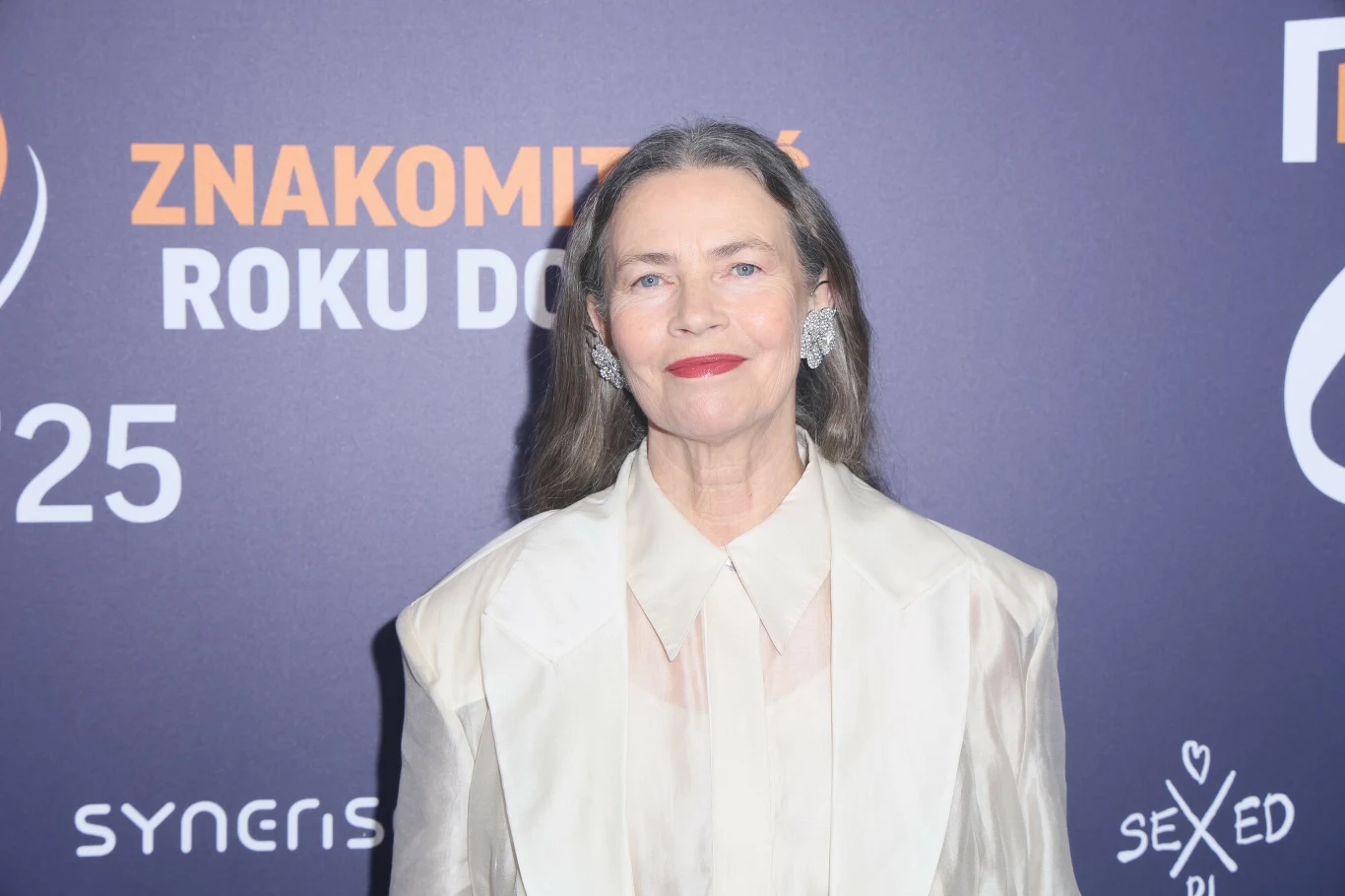 Elegancko ubrana kobieta o siwych włosach i czerwonych ustach stoi na tle ścianki z napisem 'Znakomitość roku dojrzałego', uśmiechając się delikatnie w stronę aparatu.