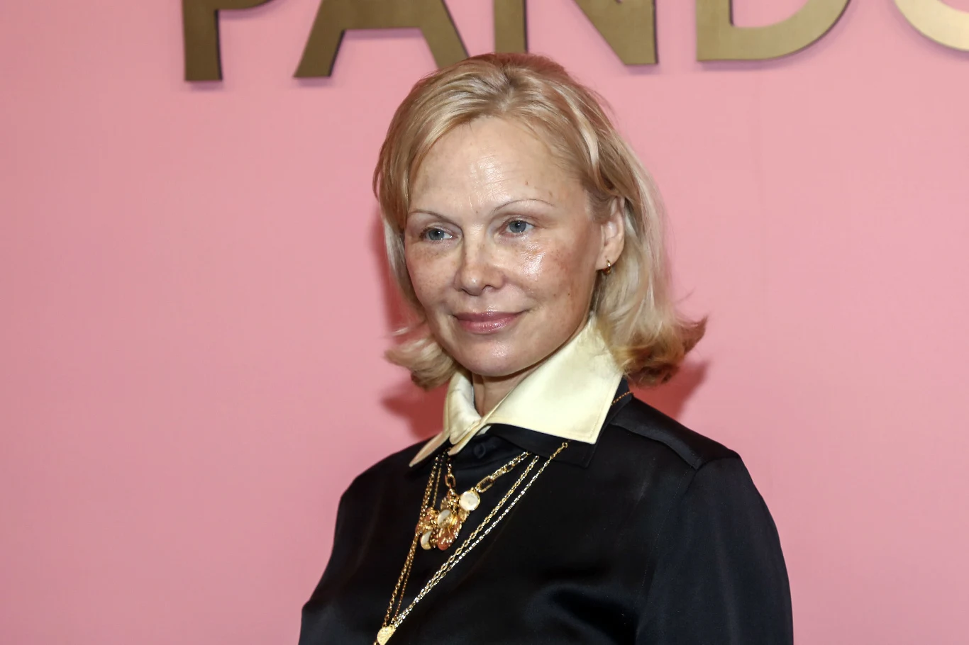 Kobieta o jasnych włosach ubrana w czarną koszulę z jasnym kołnierzem, ozdobiona kilkoma złotymi naszyjnikami, stoi na tle różowej ściany z dużym złotym napisem.