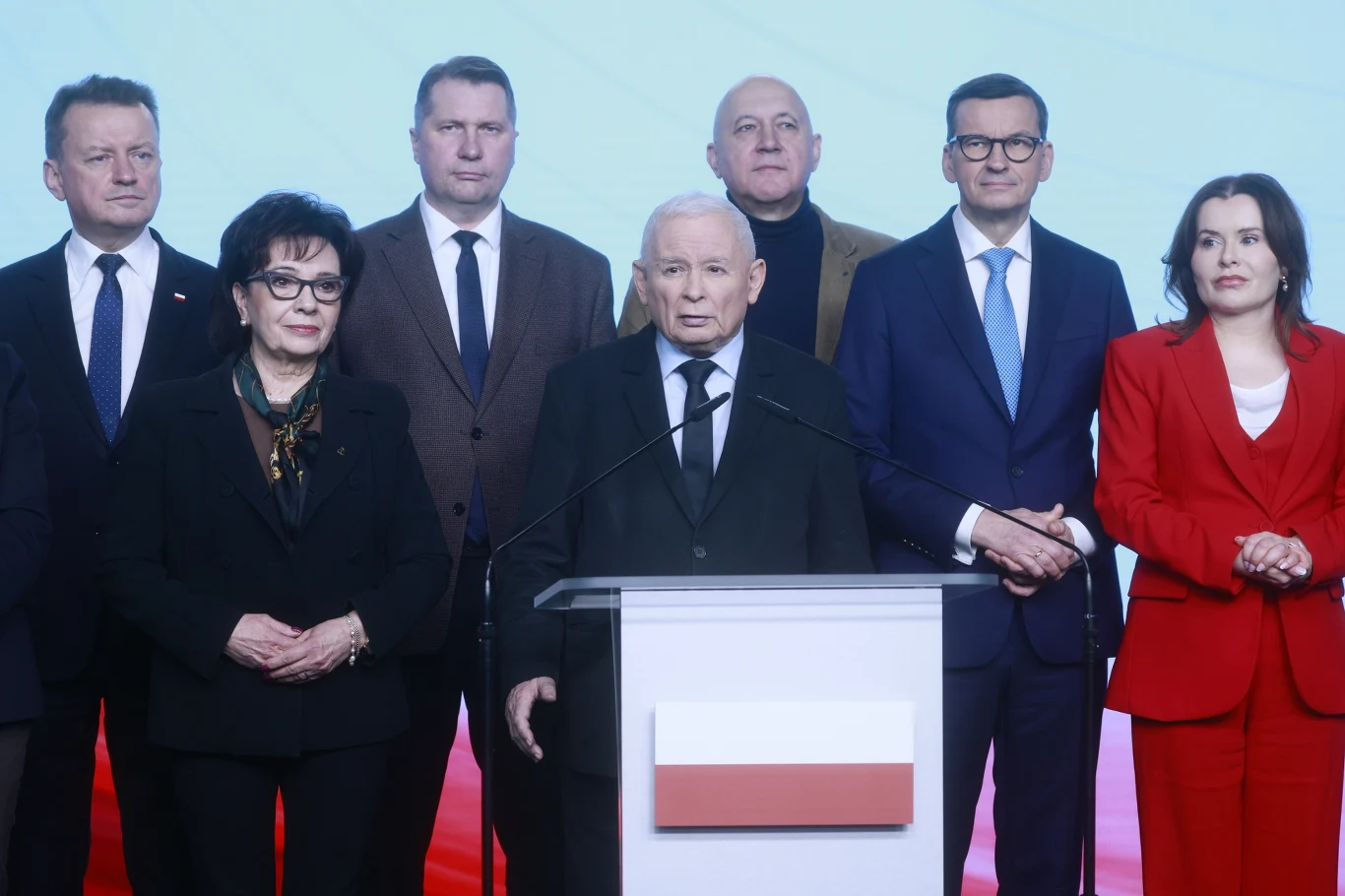 Grupa elegancko ubranych osób stoi za mównicą z polską flagą, centralny mówca jest otoczony przez innych uczestników spotkania, wszyscy skupieni na wydarzeniu.