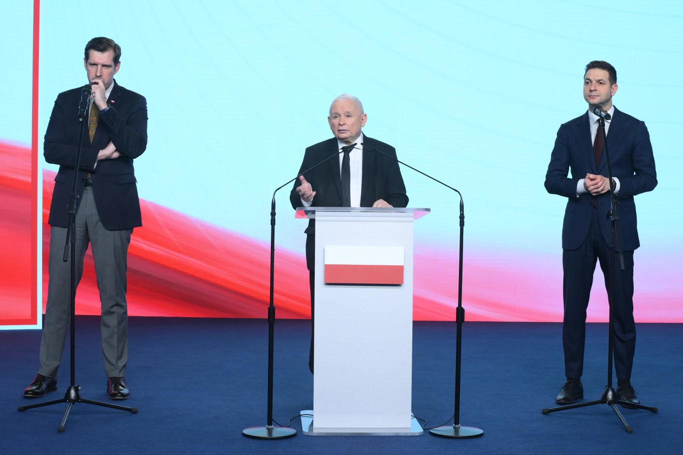Trzech mężczyzn w garniturach stoi na scenie przy mikrofonach, środkowy przemawia zza mównicy ozdobionej symbolem polskiej flagi, w tle jasne, czerwono-białe tło nawiązujące do barw narodowych.