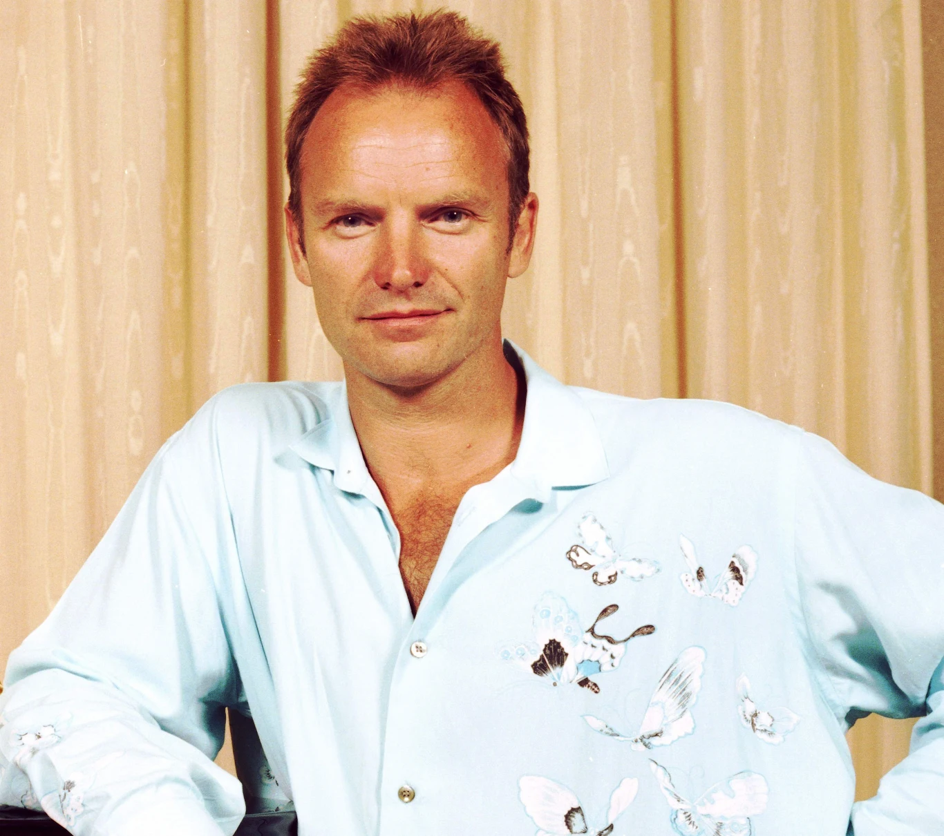 Sting w 1996 r. Sting w 1996 roku w jasnoniebieskiej koszuli z motywem motyli, stoi na tle beżowych zasłon i patrzy w obiektyw.