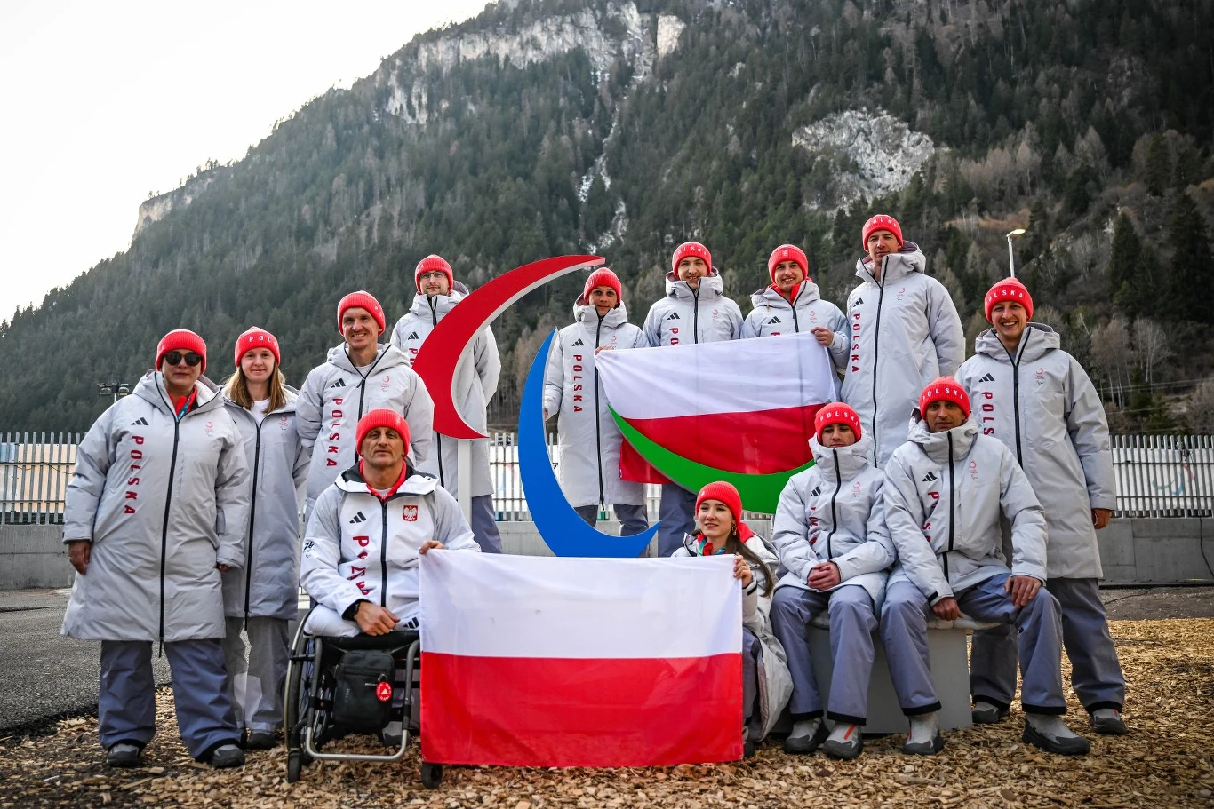 Grupa sportowców w zimowych strojach reprezentacyjnych z napisem 'POLSKA' stoi i siedzi wokół symbolu paraolimpijskiego, trzymając biało-czerwoną flagę Polski oraz flagę z zielonym, czerwonym i białym motywem na tle górskiej scenerii i ogrodzenia.