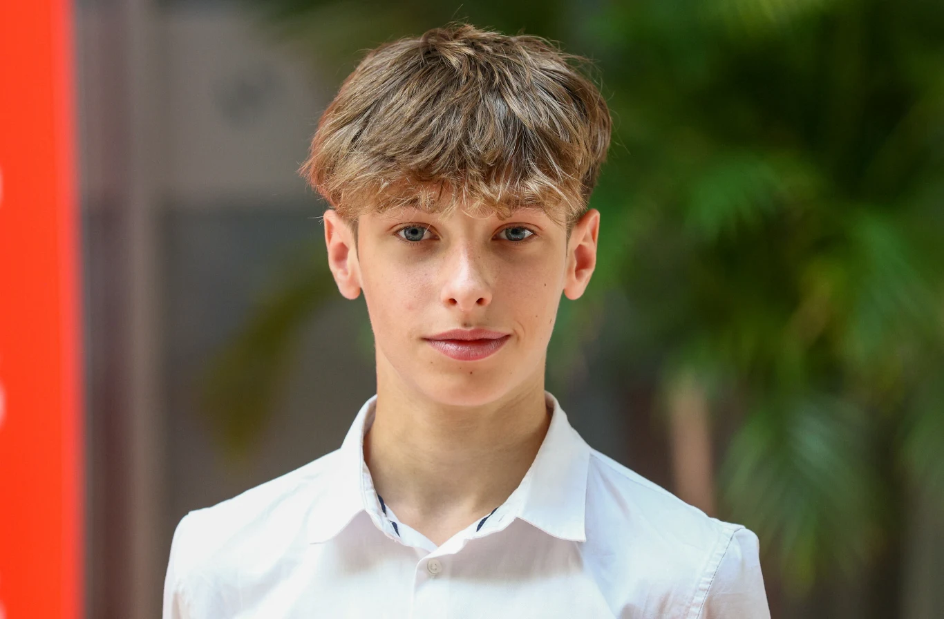 Młody chłopak o jasnych włosach i niebieskich oczach w białej koszuli, stojący przed rozmytym tłem z zielonymi roślinami.