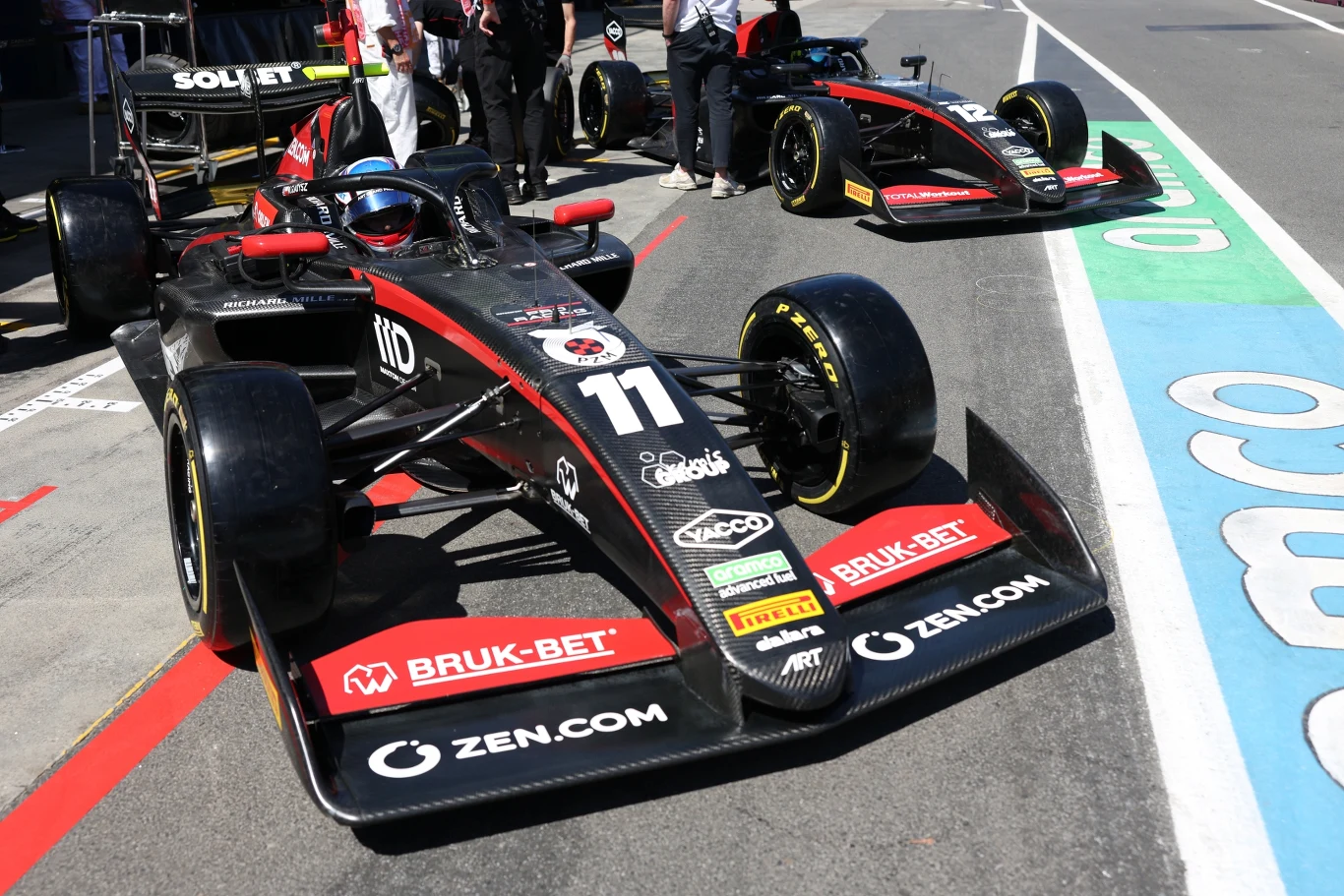Czarny bolid wyścigowy z numerem 11 na przodzie i licznymi sponsorami, takimi jak BRUK-BET i ZEN.COM, stoi na stanowisku serwisowym, a w tle widoczny jest drugi podobny bolid oraz członkowie zespołu w białych koszulkach.
