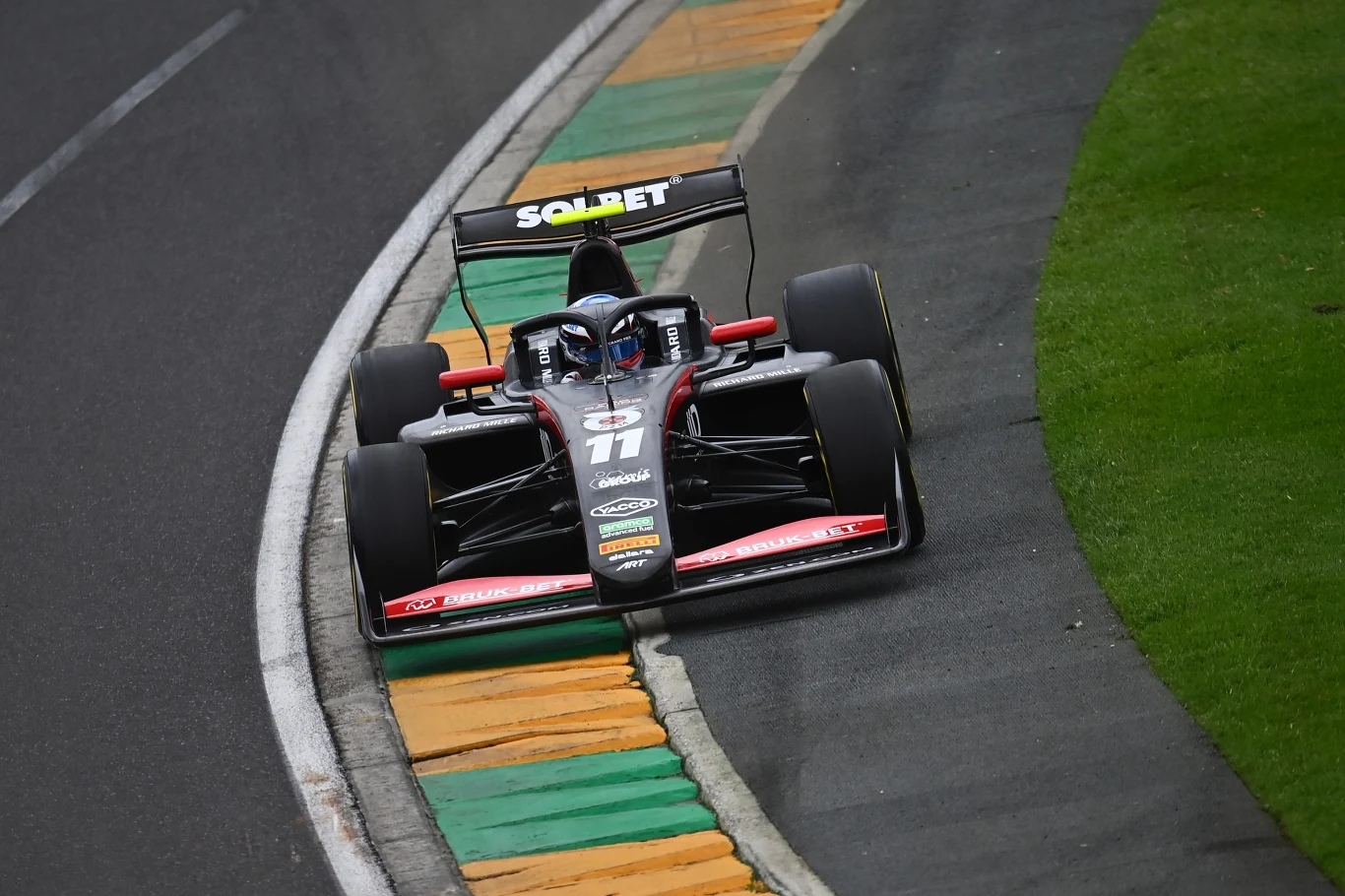 Czarny bolid wyścigowy z numerem 11 jedzie po torze, zahaczając kołem o kolorowe krawężniki, obok zielonego trawnika.