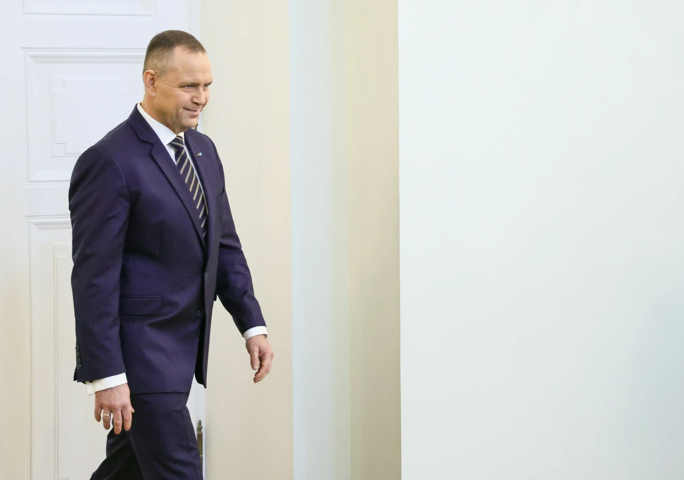 Mężczyzna w ciemnym garniturze i krawacie idzie w stronę jasnej ściany, uśmiechając się lekko.