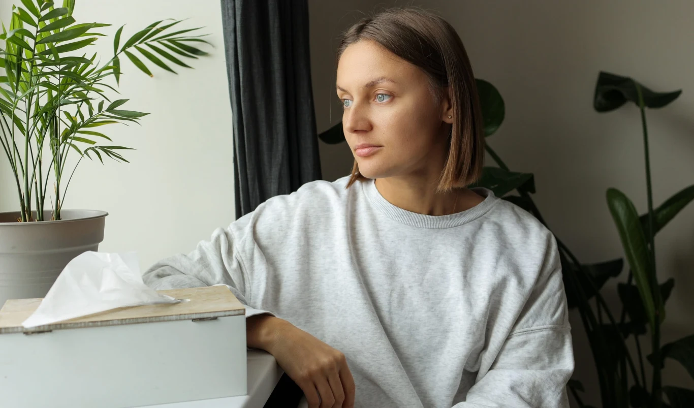 Kobieta w jasnej bluzie z krótkimi włosami siedzi przy stole, patrząc zamyślona przez okno, obok niej znajduje się pudełko chusteczek i doniczkowa roślina.