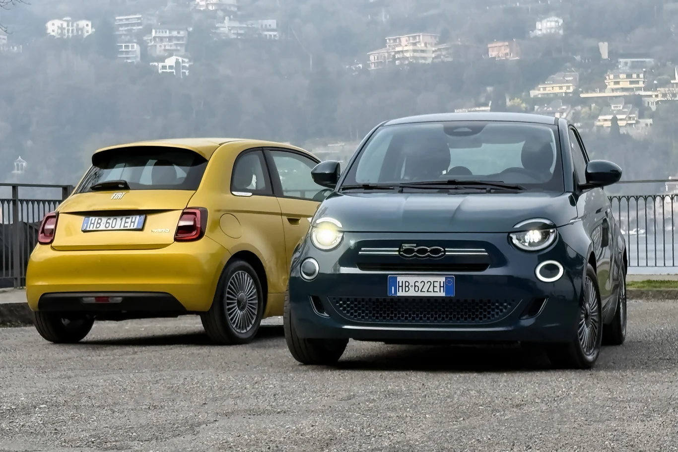 Dwa nowoczesne samochody Fiat 500, jeden w kolorze żółtym widoczny od tyłu, drugi w kolorze grafitowym widoczny od przodu, ustawione obok siebie na tle zamglonego krajobrazu miejskiego z zabudowaniami na wzgórzu.