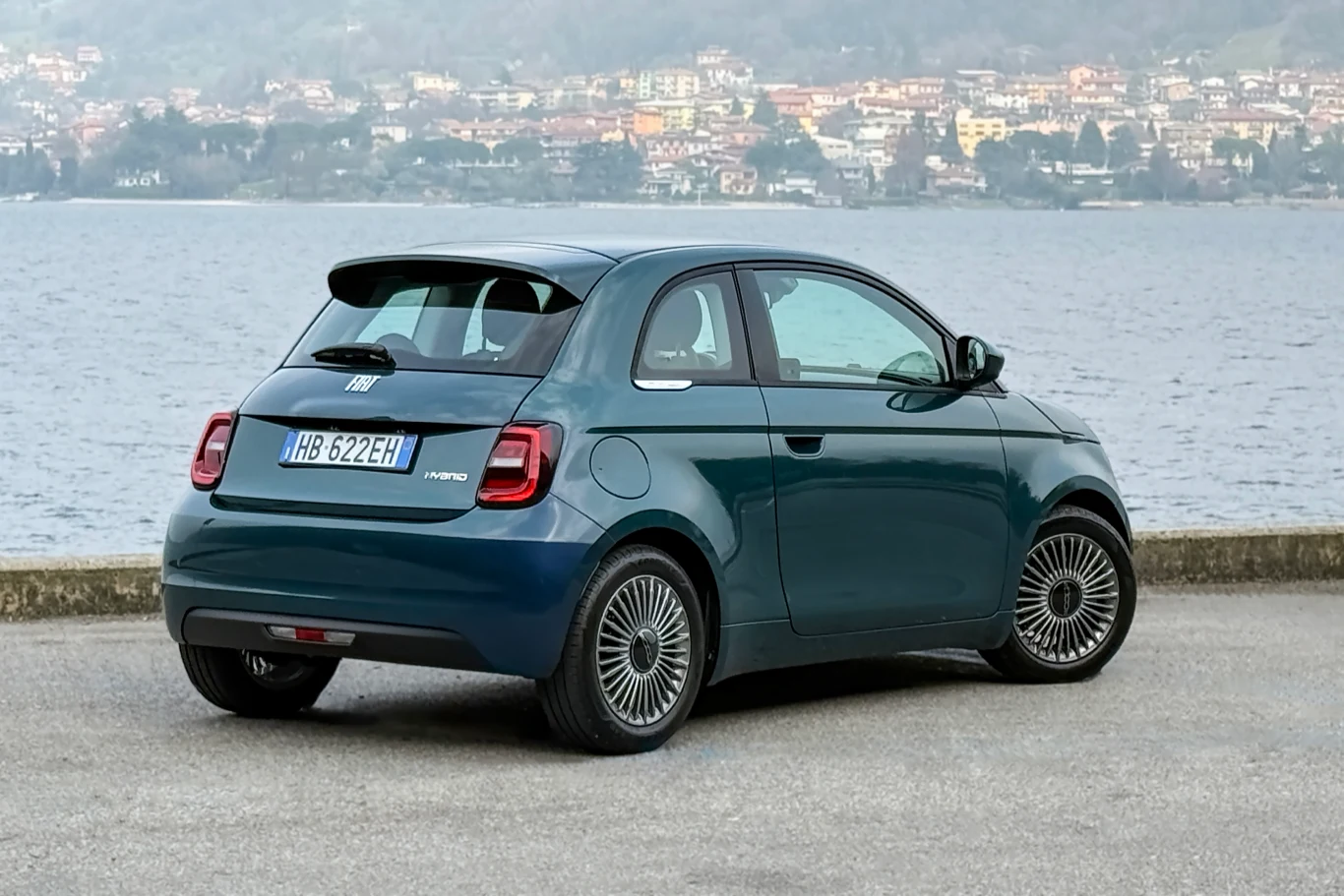 Mały, nowoczesny samochód miejski w ciemnoniebieskim kolorze zaparkowany nad brzegiem jeziora, z charakterystycznymi, stylizowanymi felgami i panoramicznymi szybami, w tle widoczna zabudowa miejska na wzgórzu.