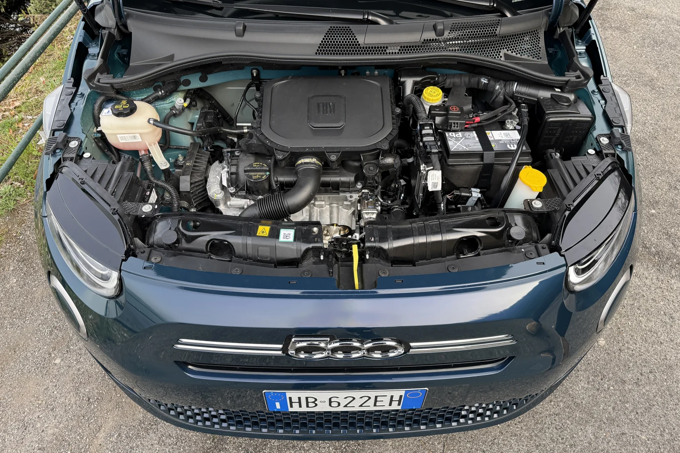 Komora silnika samochodu osobowego z widocznym akumulatorem, zbiornikiem płynu chłodzącego, elementami instalacji elektrycznej i osłoną silnika z logo producenta.