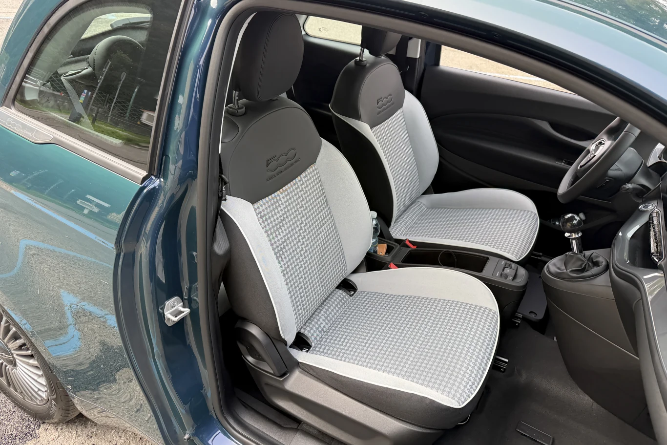 Przestronne wnętrze samochodu z dwoma przednimi fotelami pokrytymi jasnoszarą tapicerką, czarna deska rozdzielcza ze skrzynią biegów i kierownicą po prawej stronie oraz uchylone drzwi ukazujące szczegóły kabiny.