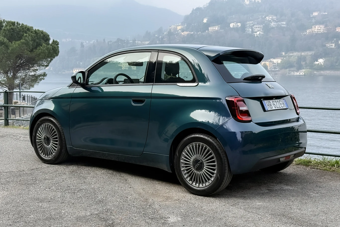 Mały, nowoczesny samochód miejski w kolorze ciemnej zieleni zaparkowany na asfaltowej nawierzchni przy metalowej barierce, w tle widok na jezioro i wzgórza z zabudowaniami.