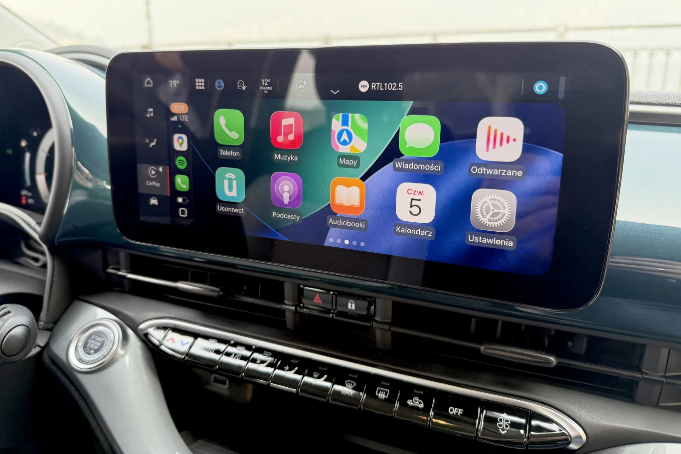 Dotykowy ekran multimedialny samochodu z uruchomionym systemem Apple CarPlay, na którym widoczne są kolorowe ikony aplikacji takich jak Telefon, Muzyka, Mapy, Wiadomości, Podcasty, Audiobooki oraz data i godzina; poniżej ekranu umieszczone są przyciski...