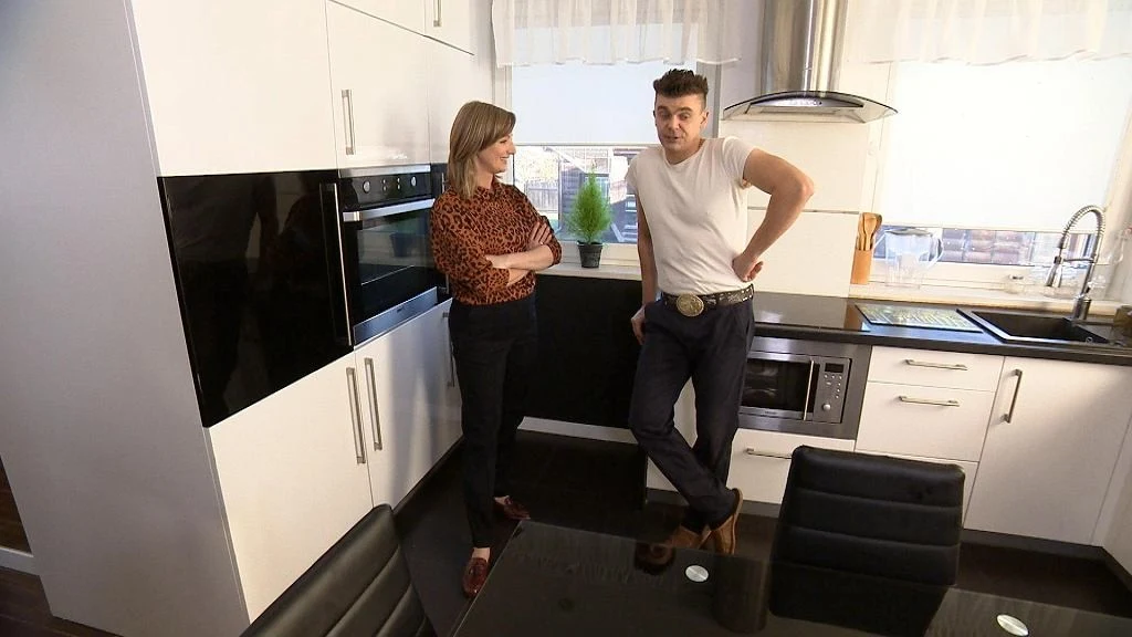 Dwie osoby rozmawiające w nowoczesnej, białej kuchni z czarnymi akcentami, przy dużym stole ze szklanym blatem, w tle okno z białą firanką i zlewozmywak.