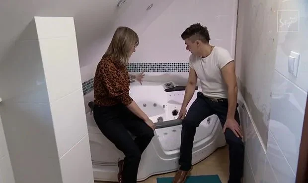 Dwie osoby siedzące na krawędzi narożnej wanny z hydromasażem w małej, jasnej łazience o skośnym suficie; jedna z nich wskazuje na elementy wanny, a obok na podłodze leży niebieski dywanik.