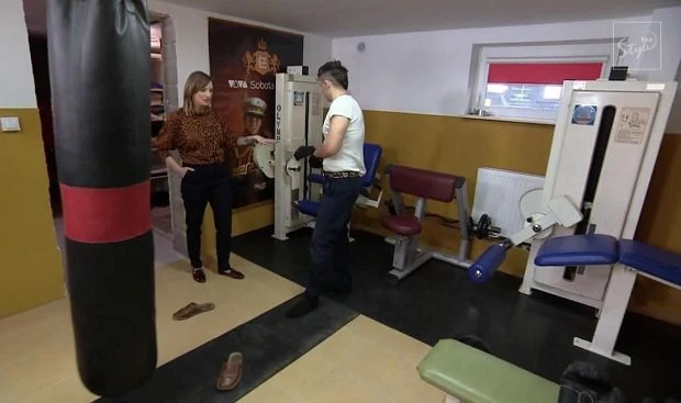 Siłownia z maszynami do ćwiczeń, workiem bokserskim oraz dwiema osobami rozmawiającymi; jedna z nich stoi boso, w tle plakat z mistrzem bokserskim.