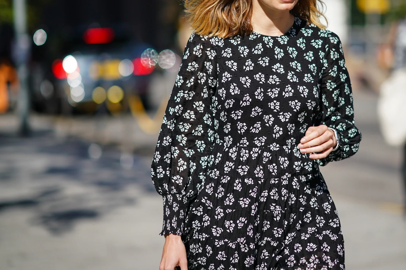 Kobieta ubrana w czarną sukienkę w drobny biały wzór kwiatowy spaceruje po mieście; widoczne delikatnie falowane włosy do ramion oraz fragment ulicy w tle.
