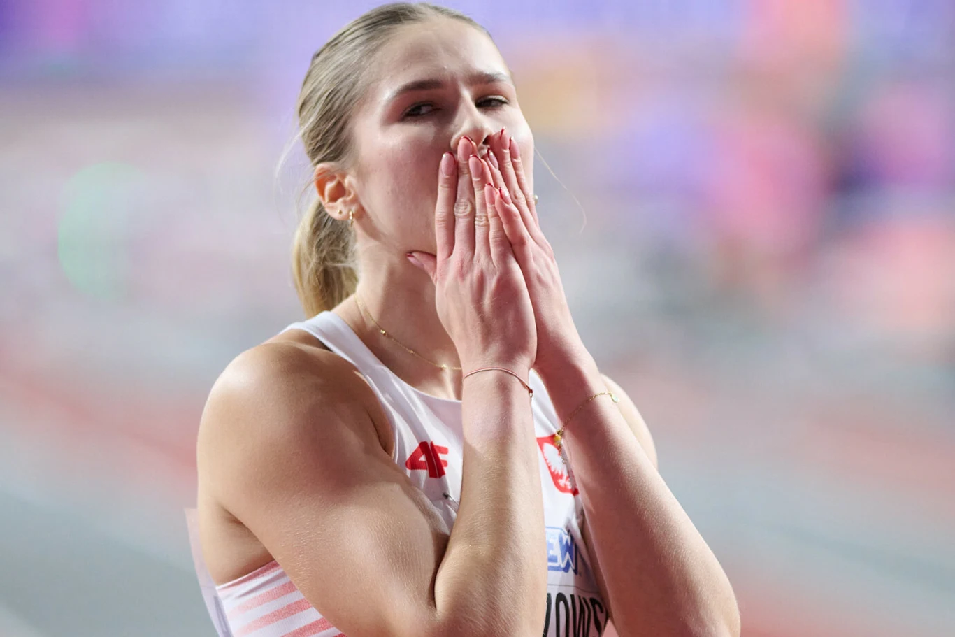 Pia Skrzyszowska Sportsmenka w biało-czerwonym stroju lekkoatletycznym z herbem Polski, stojąca na tle rozmytego stadionu, trzyma dłonie przy ustach w geście silnych emocji.