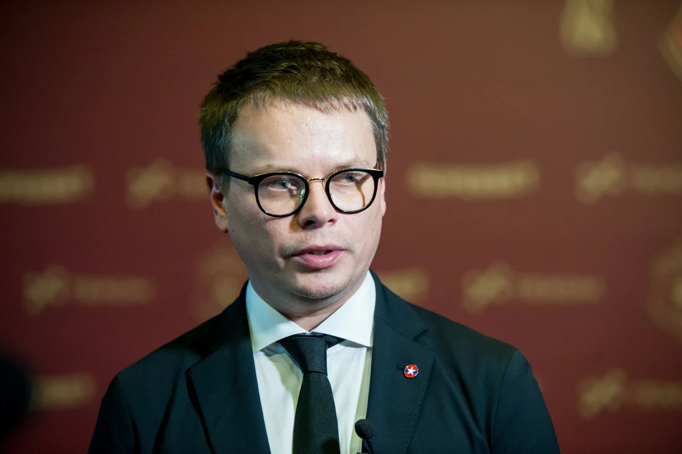 Mężczyzna w okularach ubrany w ciemny garnitur, białą koszulę i czarny krawat, trzyma mikrofon na tle bordowej ścianki z niewyraźnymi logotypami.