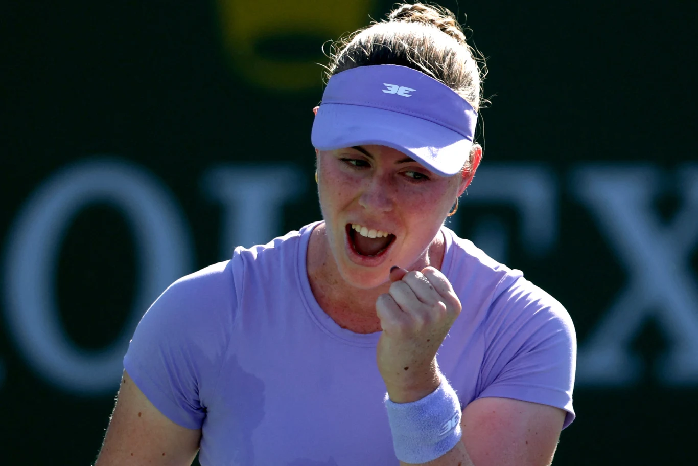 Tenisistka ubrana w fioletową koszulkę i opaskę na głowie zaciśniętą pięścią wyraża radość i determinację podczas meczu, na tle rozmazanego logotypu marki zegarków.