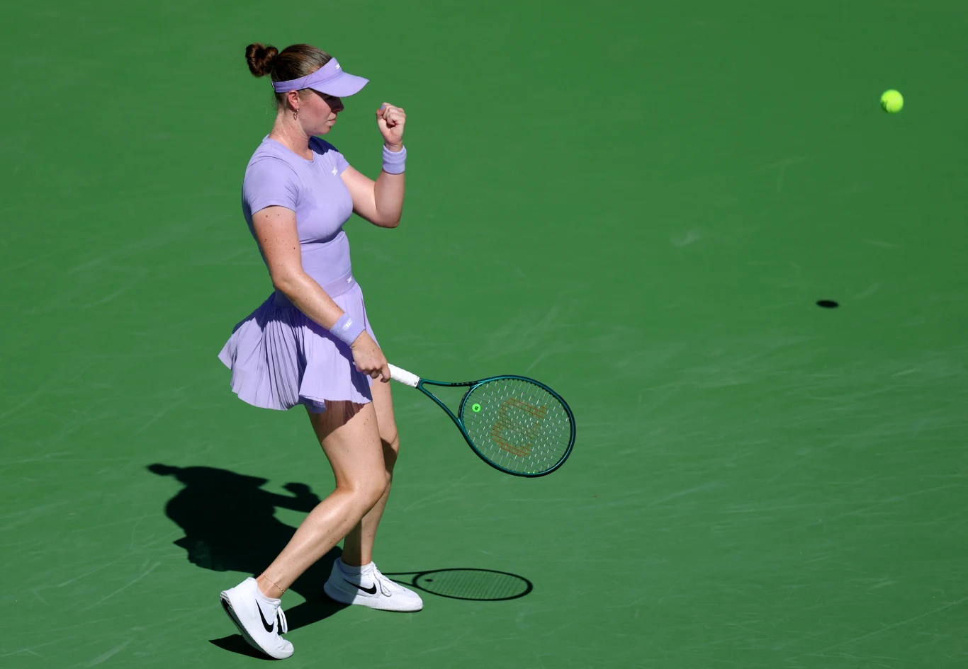 Tenisistka w fioletowej sukience i sportowej czapce świętuje udany punkt na korcie, zaciśnięta pięść wyraża emocje, w tle widoczna żółta piłka tenisowa.