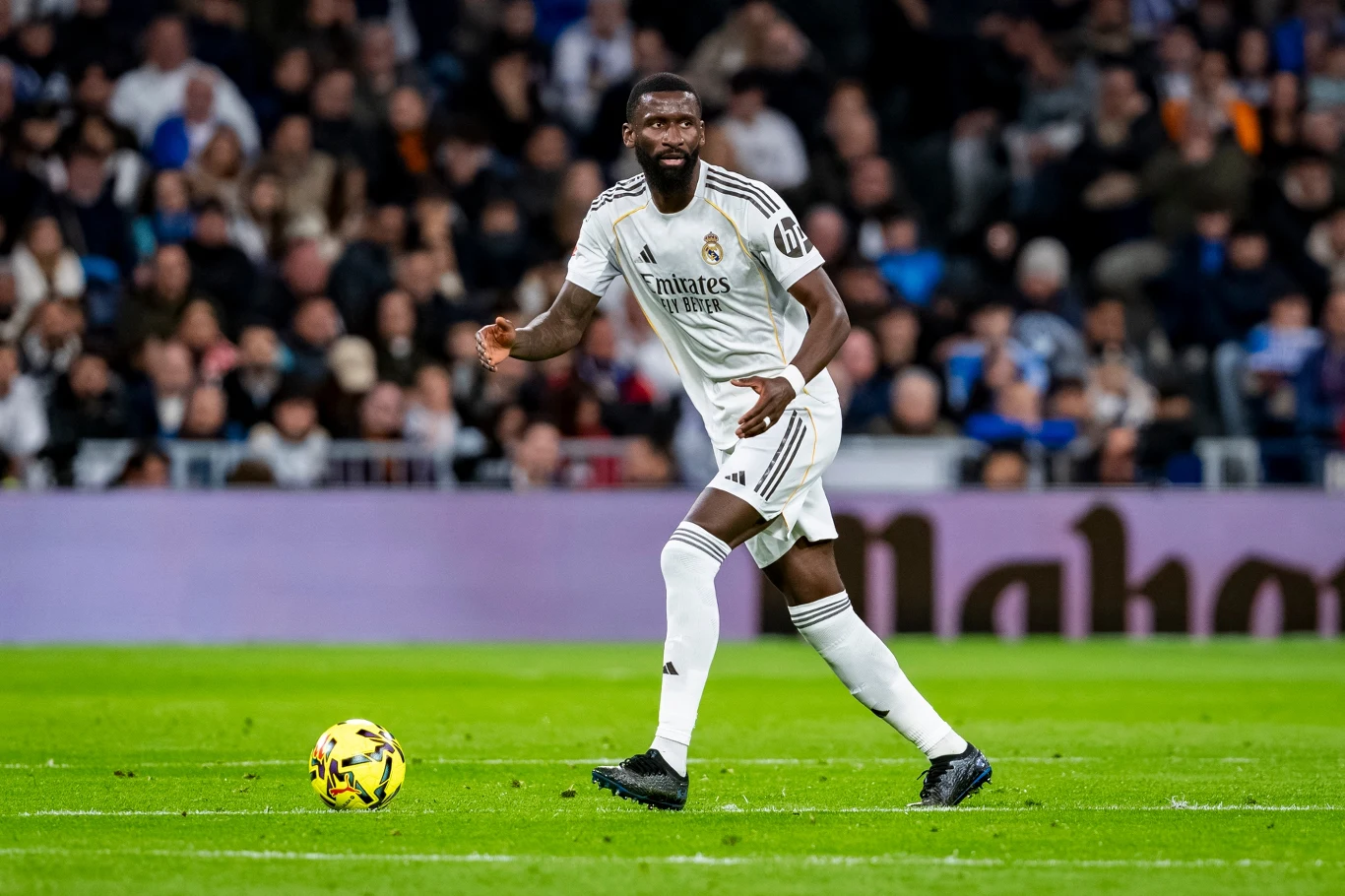 Piłkarz w białym stroju Realu Madryt kontroluje piłkę na boisku podczas meczu, w tle widoczna trybuna z kibicami.