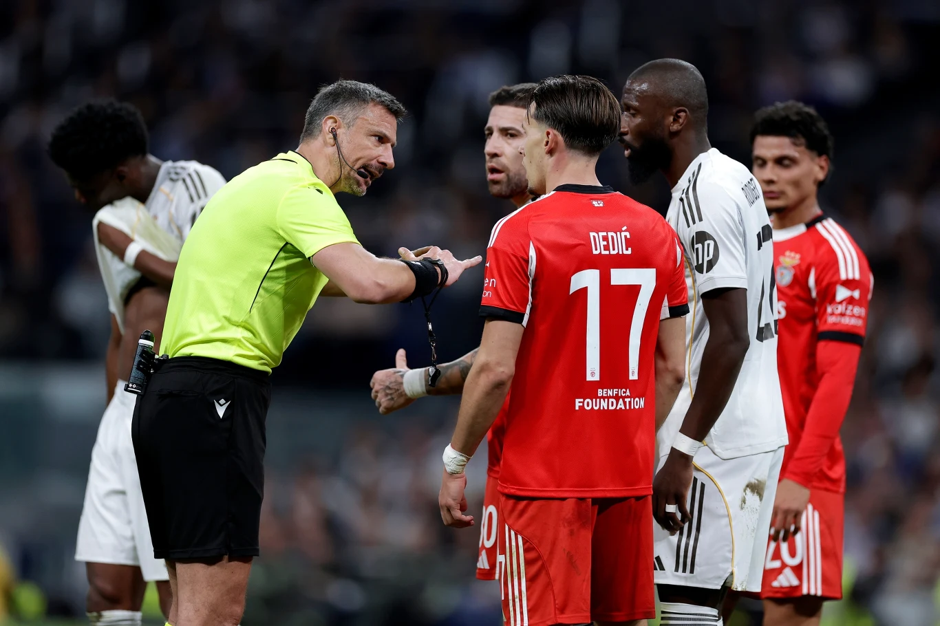 Sędzia w żółtej koszulce dyskutuje z piłkarzami obu drużyn podczas meczu, wskazując coś ręką; w centrum uwagi znajdują się zawodnicy w czarno-białych oraz czerwonych strojach, a atmosfera jest napięta.