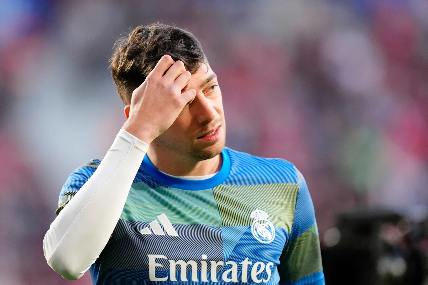 Piłkarz w stroju treningowym Realu Madryt trzyma się za głowę, sprawiając wrażenie zmartwionego lub zamyślonego, na tle rozmytego stadionu.