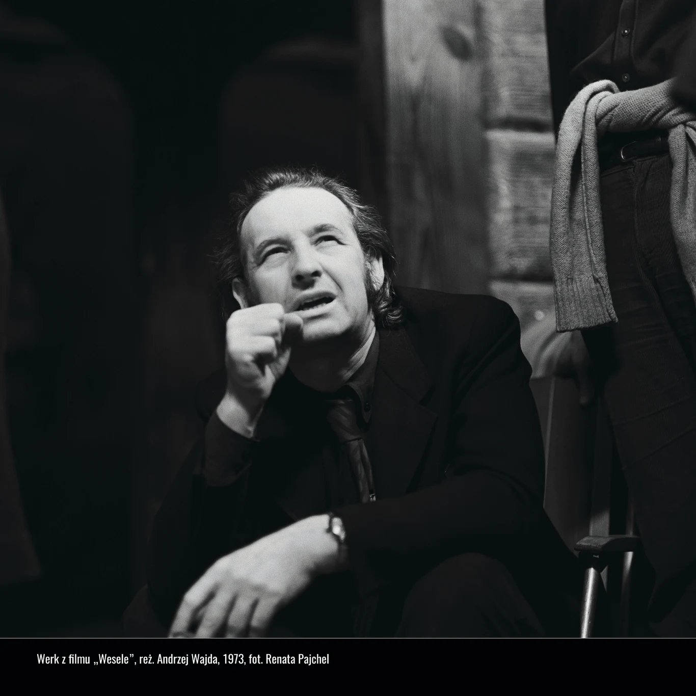 Mężczyzna w średnim wieku siedzący w ciemnym pomieszczeniu, z zamyśloną miną patrzy w górę, wspierając brodę na dłoni; w tle fragment drewnianej ściany oraz inna postać z przewieszonym przez ramiona swetrem.