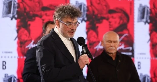 Mężczyzna z siwymi, kręconymi włosami i brodą, ubrany w elegancki garnitur, mówi do mikrofonu, a za nim stoją dwaj starsi mężczyźni w czarnych marynarkach; w tle wyraźna czerwona grafika z podobizną osoby i znaczkiem pocztowym.