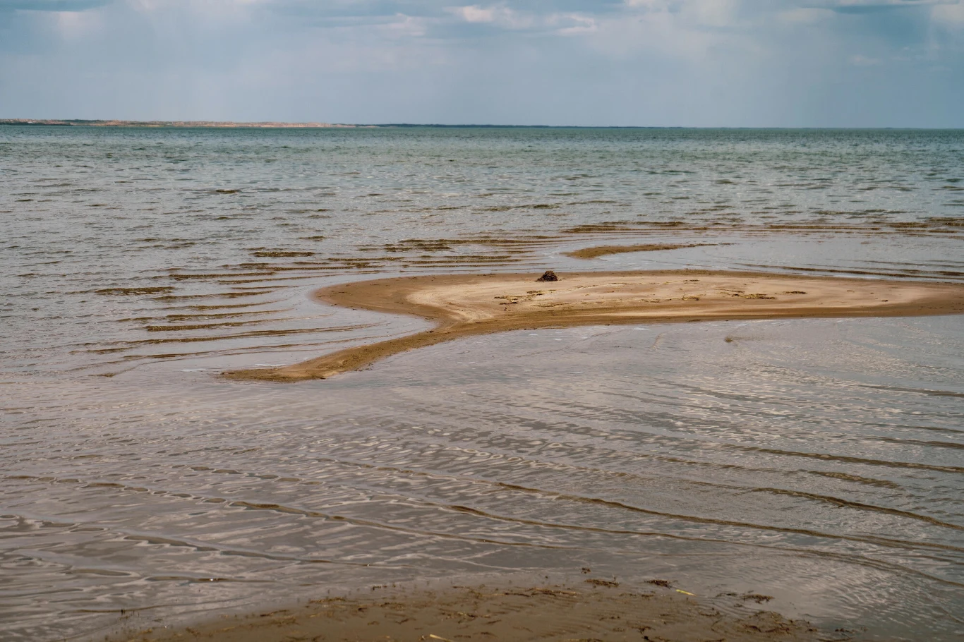 Piaszczysta łacha otoczona płytką wodą, pośrodku której leży pojedynczy ciemny przedmiot, w tle spokojne morze pod pochmurnym niebem.