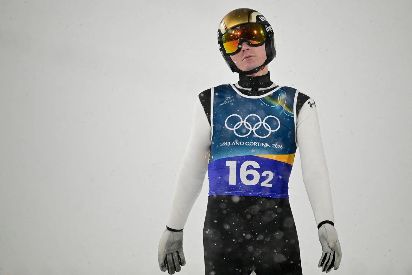 Skoczek narciarski w kombinezonie i kasku z lustrzanymi goglami, stojący na tle śniegu podczas zawodów olimpijskich w Mediolanie-Cortinie 2026, z widocznym numerem 162 na piersi i olimpijskimi pierścieniami na plastronie.