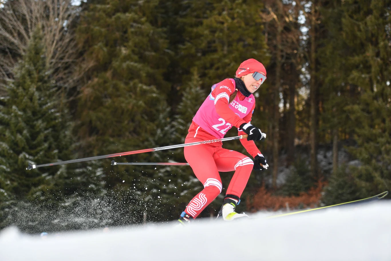 Jewgienija Krupicka Narciarz biegowy w czerwonym stroju i kasku z numerem 22 dynamicznie pokonuje trasę w lesie, energicznie odpychając się kijkami na śniegu.