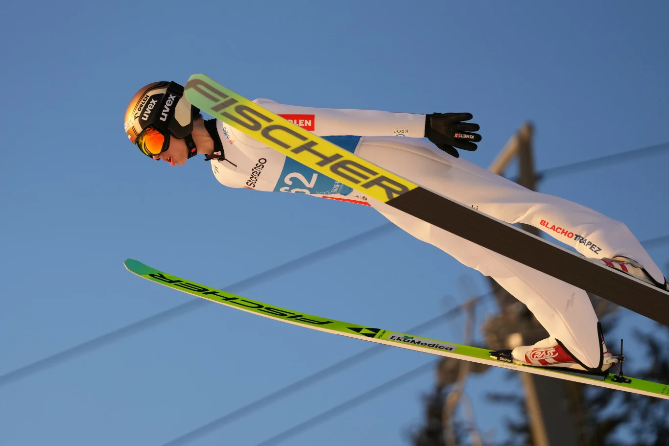 Skoczek narciarski w białym kombinezonie i kasku w trakcie lotu na nartach podczas zawodów, tło stanowi czyste niebo i niewyraźne elementy infrastruktury skoczni.