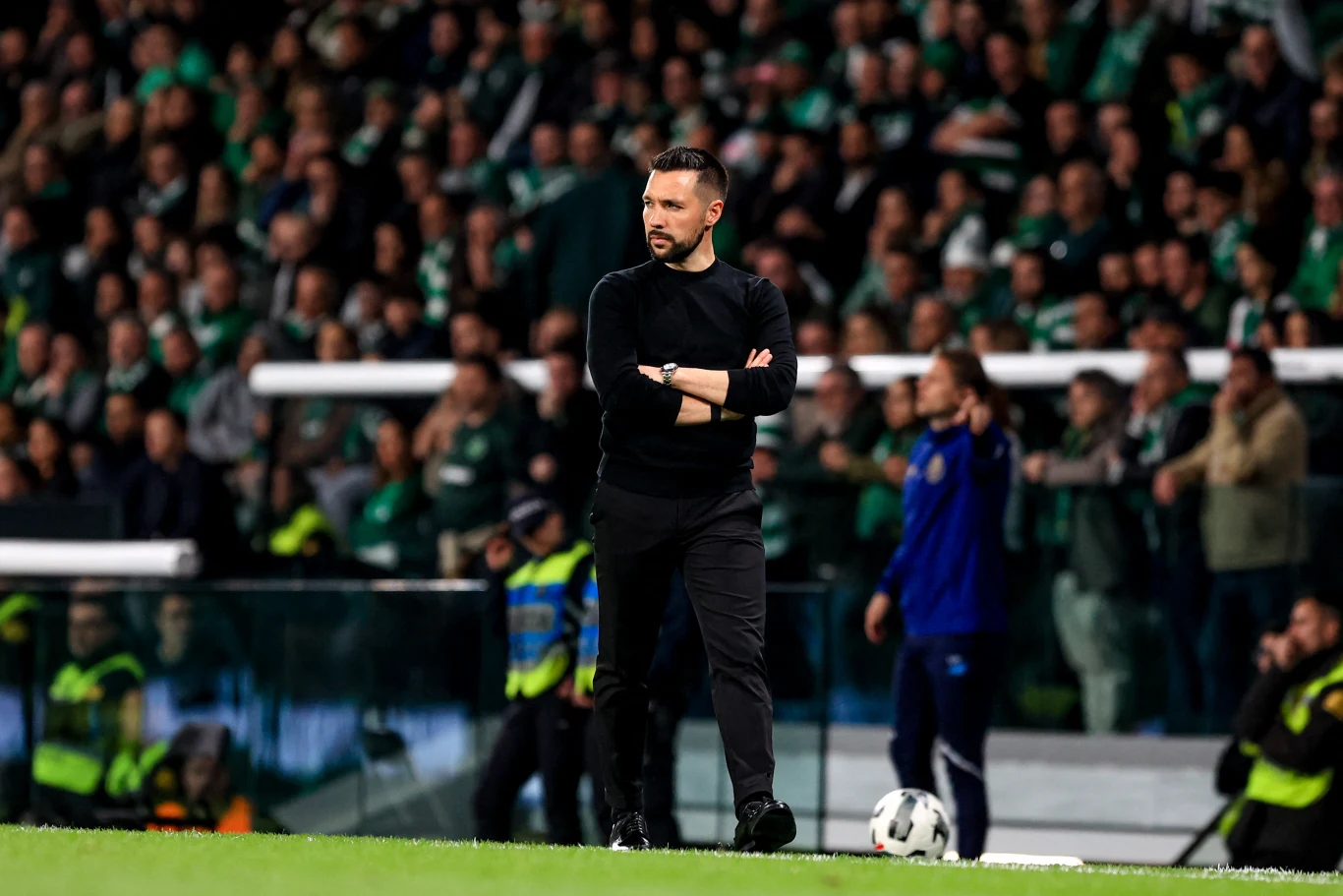 Trener w czarnym stroju stoi z założonymi rękami przy linii bocznej boiska, skupiony na przebiegu meczu, w tle tłum kibiców w zielonych barwach oraz służby porządkowe.