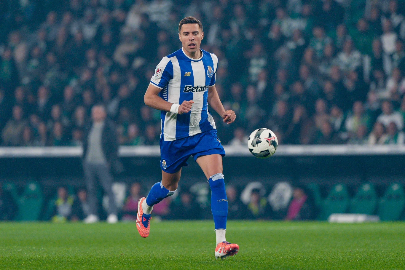 Piłkarz w niebiesko-białej koszulce i niebieskich spodenkach biegnie z piłką po zielonej murawie stadionu podczas meczu, na trybunach widać skupionych kibiców.