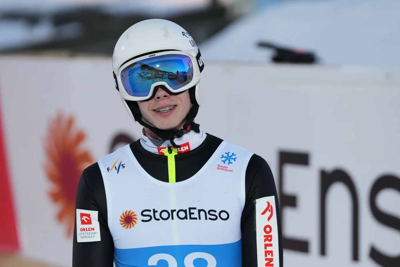 Młody skoczek narciarski w kombinezonie sportowym, kasku i goglach, z numerem startowym oraz logotypami sponsorów na tle bandy reklamowej Stora Enso.